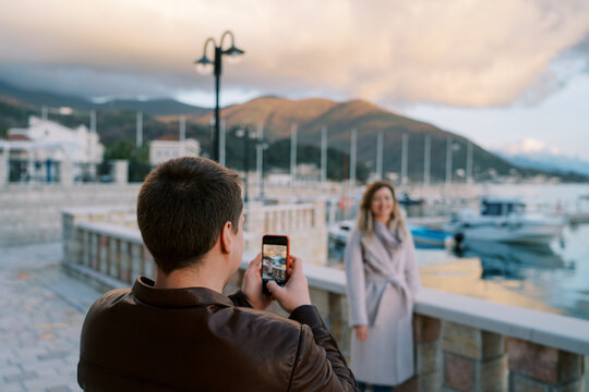 Man takes a picture with his phone of woman at the fence on the pier with moored boats. Back view