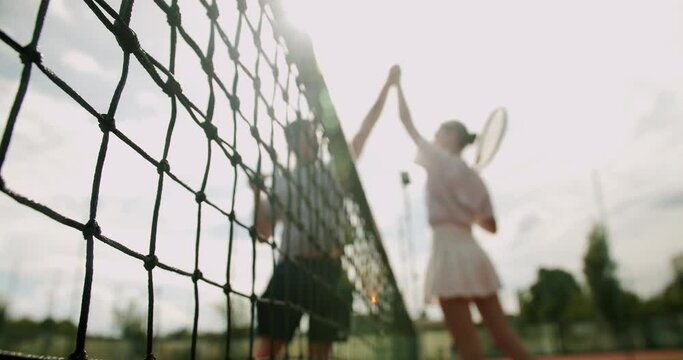 Closeup on tennis net and players woman with man on background giving high five on court after success match. Teamwork concept. Young active couple spend time together and play tennis outdoors
