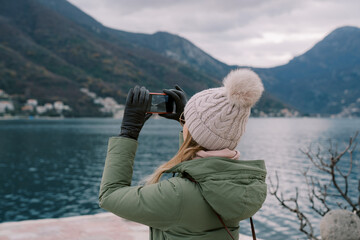 Young woman shoots mountains on the opposite shore with a smartphone from the seashore. Back view