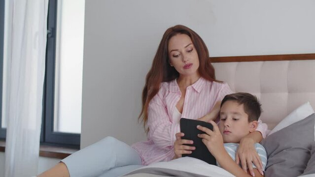 A boy lies on the bed and gets ready to sleep, reading an ebook on a tablet. A young and caring mother sits next to son and strokes his head with careness. The concept of responsiveness in the family.