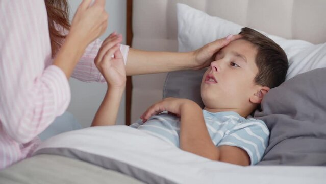 An ill boy feel bad, blows nose and wipes it into a disposable tissue, lies in the bedroom on the bed. A caring young mother sits nearby and supports the son, strokes and touches his forehead with