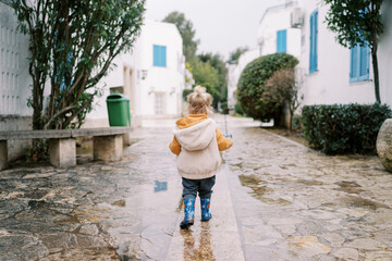 Fototapeta na wymiar Little girl walks through the puddles in the courtyard. Back view