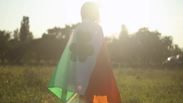Little girl covered with large flag of Ireland walking outdoors in bright sun on St Patrick's Day