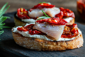 Two bread slices with ricotta cheese, pieces of herring and sun dried red tomatoes on wooden board...