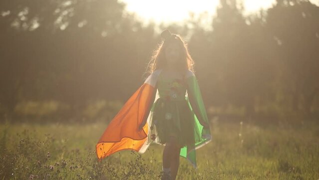 Cute redhead girl dressed as a green fairy walking outdoors with flag of Ireland on shoulders. Irish child celebrating St Patricks Day