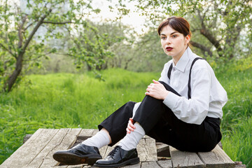 A beautiful girl in a white shirt, in black trousers with suspenders against the background of the sky and green grass