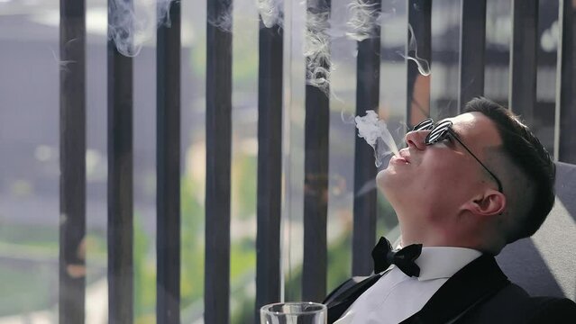 An elegant man in a black suit and black glasses sits on the balcony, holds a glass of alcohol in his hands, smokes and exhales cigarette smoke. Preparation of the groom for the difficult wedding day.