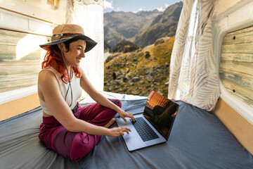 Red-haired woman teleworking typing with laptop in camper van in middle of nature near lake, connected to internet, girl writing with laptop, smiling showing computer