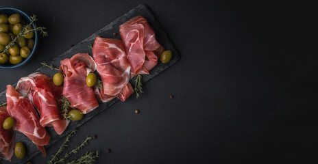 Appetizer from dry cured serrano ham or Spanish jamon iberico. Italian prosciutto crudo served with green olives on the black slate stone board. Meat slices on the black background - obrazy, fototapety, plakaty