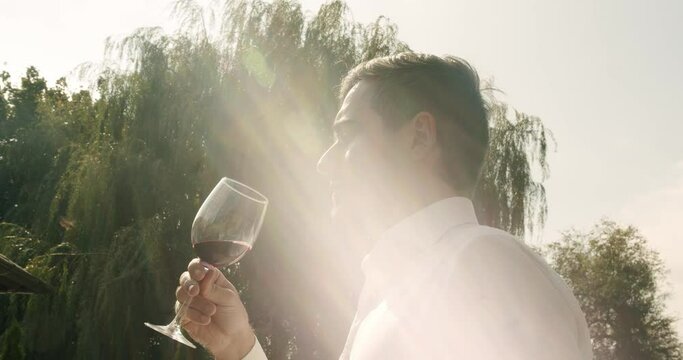 Man sommelier in white shirt hold glass with red wine in hand, sniffing flavor and tasting drink outdoors at sunny day. male expert appreciating wine