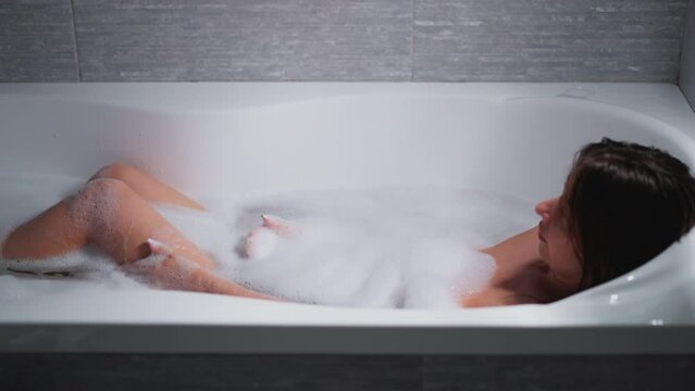 Woman strokes legs taking bath in bathtub at home. Lady washes elegant body during spa procedure in bathroom. Woman pampers soft skin in soap water
