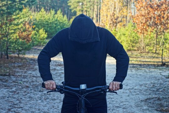 one man dressed in a black fashionable stylish raglan and a hood on his head stands on the road and holds the steering wheel along with a black modern new mountain iron bike in the forest on vacation 