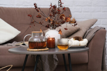 Autumn home design - hot teapot with tea, glass of tea, burning candle, vase with dry branches on sofa background in cozy living room.