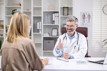 Fototapeta premium Back view, experienced mature doctor consulting smiling female patient, gray-haired man working inside clinic office, using laptop.