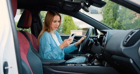 Beautiful young woman driver in smart clothes using mobile phone while sitting in luxury car. Young girl sitting behind wheel of premium modern car with smartphone in her hands.