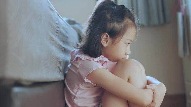 Adorable Asian kid girl is sitting on the bedroom floor alone. Her face thoughtfully expresses anxiety, sadness, sorrow and depression in her eyes and face with lonely and exhausted posture.