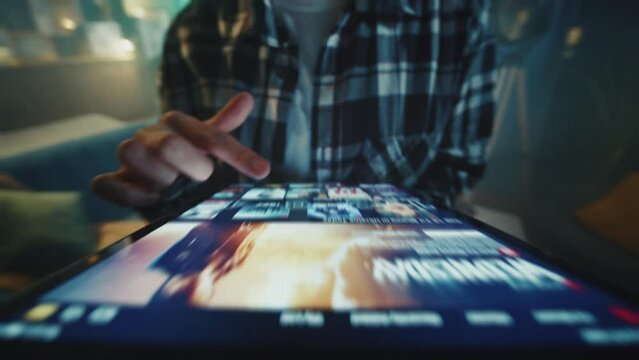 Shot of female hipster wearing checkered shirt holding digital tablet in hands. Young unrecognizable woman choosing TV show to watch on streaming platform. Blurred background of living room. Indoors