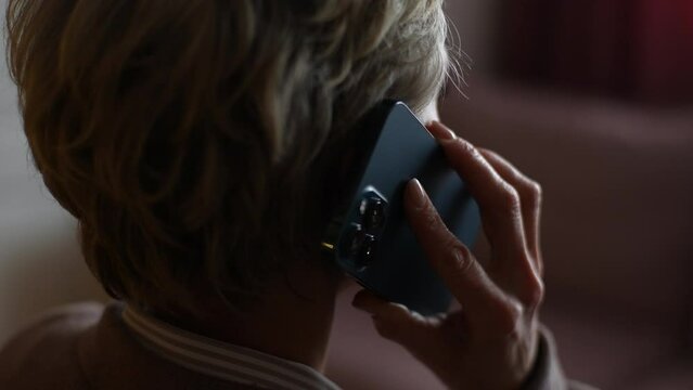 Closeup back view of unrecognizable middle-aged woman answering phone, brings smartphone to ear in dark room. Close-up rear view of elderly female chatting sharing news on mobile phone, slow motion.