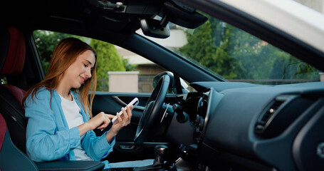 Naklejka premium Cheerful blonde girl with freckles sitting in modern car and using mobile phone for surfing internet social media e-mail. Woman using touch screen in car setting up the navigation gps application on