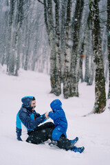 Fototapeta na wymiar Laughing dad holds the hands of a small child sitting on his lap during a snowfall in the forest