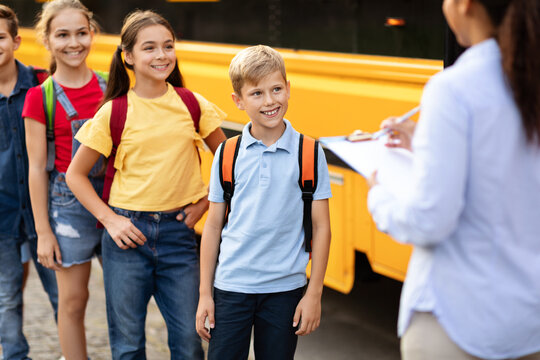 Black female teacher checking off names as children board the school bus