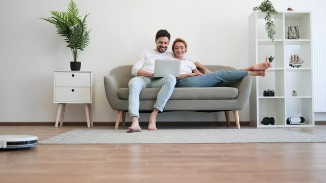 Young caucasian couple in casual clothes relaxing on comfy couch with portable laptop in hands. Family of two watching movie while wireless robot vacuuming wooden floor.