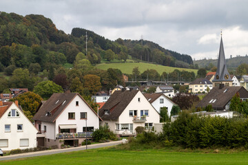 Fototapeta na wymiar village in the mountains, images shows a small village in germany near the town of paderborn, consisting of few houses, shops and a church with spire, with the steep hills and trees in the background