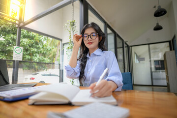 Asian businesswoman wearing glasses sits at her desk. Focus on writing in notebooks.