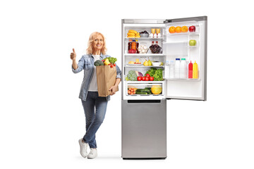 Mature woman with a grocery bag leaning on an open fridge and gesturing thumbs up