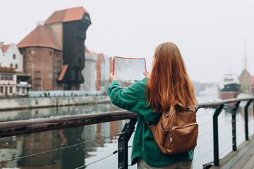 Attractive young female tourist is exploring new city. Redhead girl with backpack holding a paper map on city street in Gdansk. Traveling Europe in autumn. Famous Zuraw crane, Motlawa river
