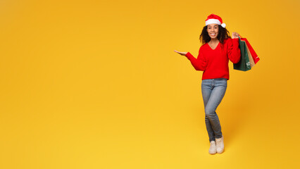 Black lady in Santa hat with bags, pointing to space, yellow background, full length