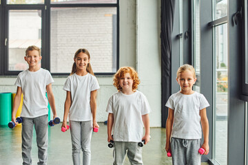 Obraz premium four cheerful little children posing with dumbbells and smiling joyfully at camera, child sport