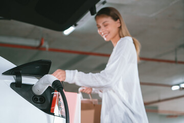 Young woman travel with EV electric car to shopping center parking lot charging in downtown city...
