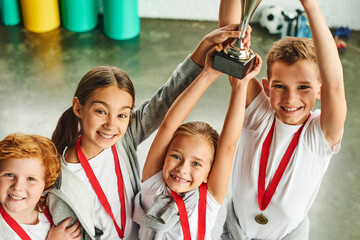 four cheerful cute preadolescent children in sportswear triumphing with huge trophy in hands