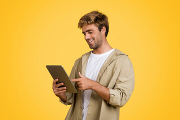 Joyful European man surfing web on tablet, yellow backdrop