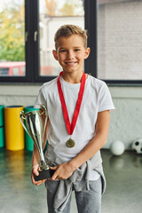 vertical shot of little cute boy with golden medal and trophy smiling at camera, child sport