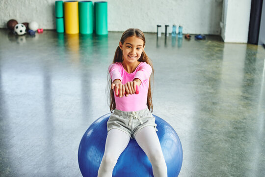 preadolescent cute girl in pink sportswear exercising with dumbbell on fitness ball, child sport