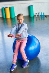pretty little girl in sportswear sitting on fitness ball and exercising with dumbbells, child sport