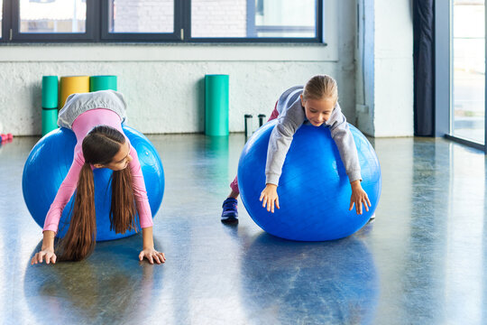 two joyful girls in sportswear lying on fitness balls in gym stretching hands to floor, child sport