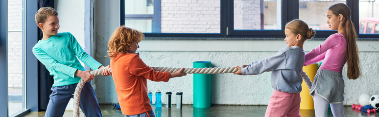 four pretty children in sportswear playing tug of war with fitness rope in gym, child sport, banner