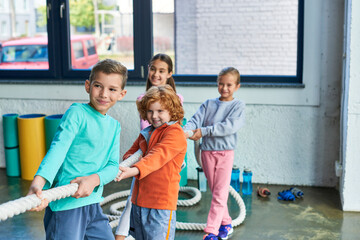joyous pretty children in sportswear pulling fitness rope in gym and smiling cheerfully, child sport