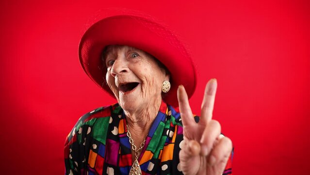 Happy fisheye portrait caricature of funny elderly woman with red hat giving peace sign gesture isolated on red background.