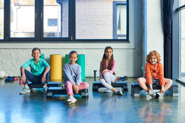 four preadolescent boys and girls sitting on fitness steppers and smiling at camera, child sport