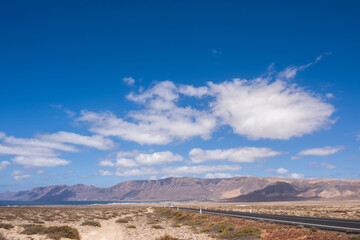 Obraz premium Desert landscape of white sand and desert bushes. Ocean and Famara cliff in the background. Dirt track. Sky with big white clouds. Lanzarote, Canary Islands, Spain.