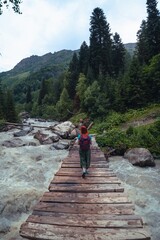 A suspension bridge on hiking trail through green dense forest with a female traveler. Woman tourist standing on wooden bridge over a raging Dolra river, Georgia. Vertical photo