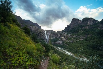 Fototapeta premium An incredible view of a majestic Shdugra waterfall in the distance, high in the Caucasus Mountains, rocky, a rushing stream. Green herbs and plants with flowers. Mazeri, Svaneti, Georgia