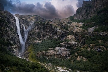 Fototapeta na wymiar An incredible view of a majestic Shdugra waterfall, high in the Caucasus Mountains, rocky slopes, a rushing stream. Mazeri, Svaneti, Georgia. Dramatic clouds before rain at sunset