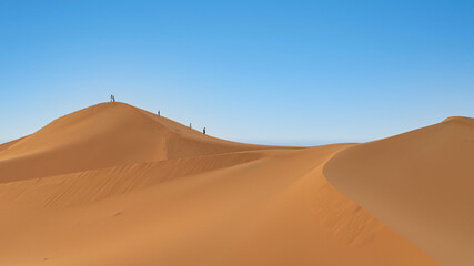 Unidentified people walking on sand dunes of Sahara Desert in northern Africa, a vast expanse of sand and dunes that stretches as far as the eye can see.