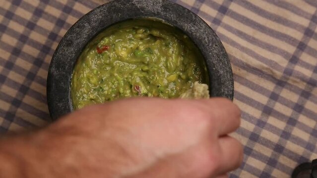 dipping a tortilla chip into guacamole (spicy mexican avocado dip served in a molcajete or stone mortar and pestle) cilantro, onion, tomato, jalapeno pepper (man's hand, caucasian model)