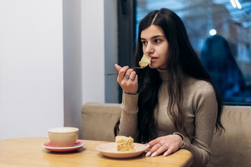 Pretty young woman sitting in cafe, drink cup of coffee hot beverage, eating piece of creamy chocolate cake with nuts on plate, tasting dessert. Chill, relax, lunch 
